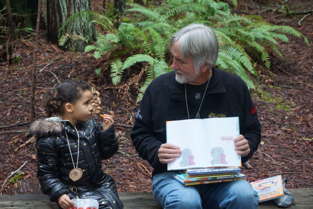 little girl sits underneath redwoods listening to a book red by a landpaths volunteer
