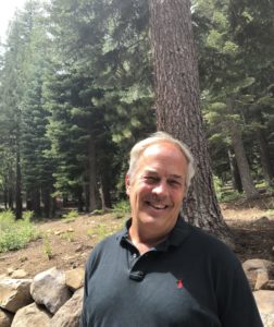 Board member Tim Choate in a black t-shirt is smiling in the trees.