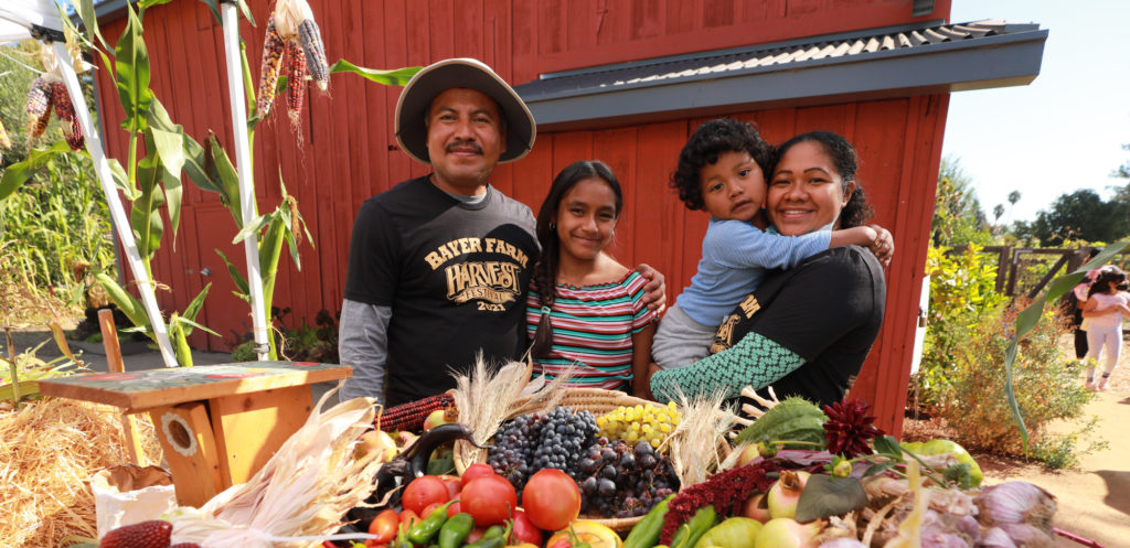 A family smiles with the red barn at Bayer Farm in the background. In the foreground of the photo is a table full of tomatoes, squash, corn, and other vegetables harvested at Bayer Farm that day.