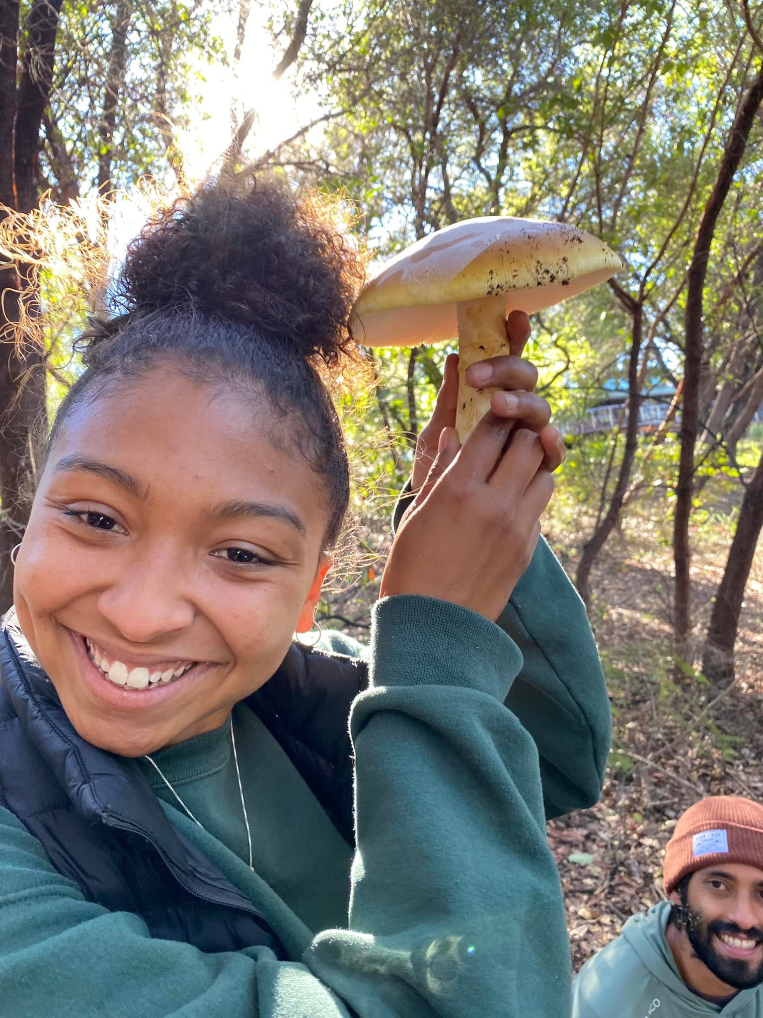 A person is smiling and holding a large mushroom up above their head with sun shining through trees in background.