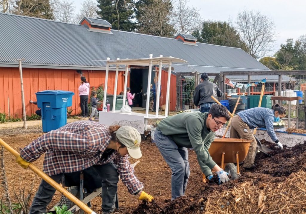 Three people scoop compost into wheelbarrows in front of a large red barn at a community garden.