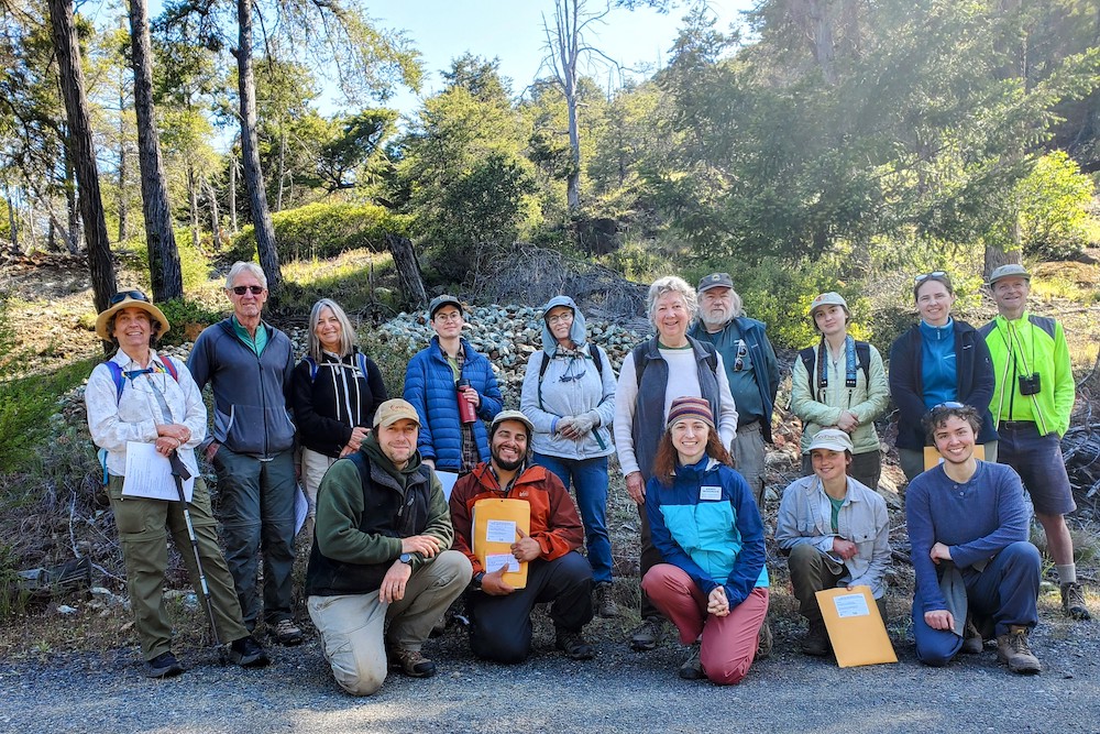 10 people stand and smile, holding folders and clipboards in front of a forest at a LandPaths' preserve. 