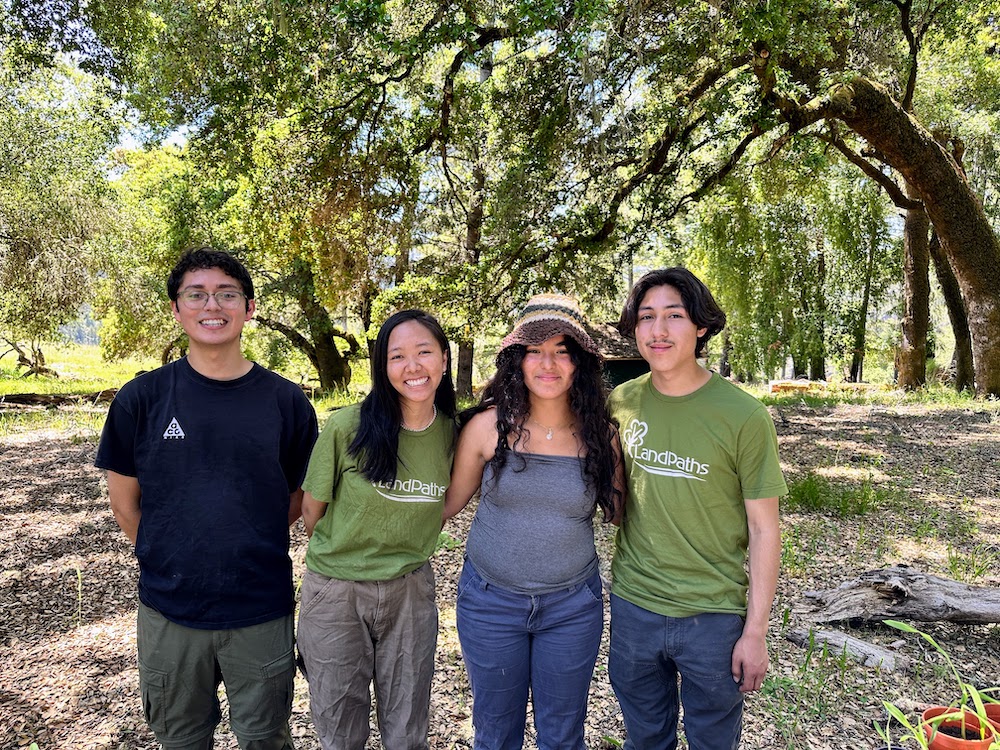 For young people stand in front of a stand of oak trees. They are smiling and look happy.