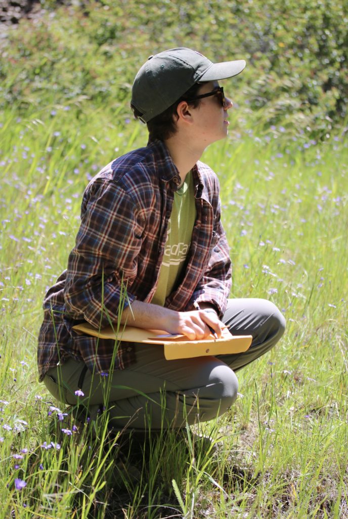 A person sits in a field of grass, holding a notebook and look outside of the frame. 