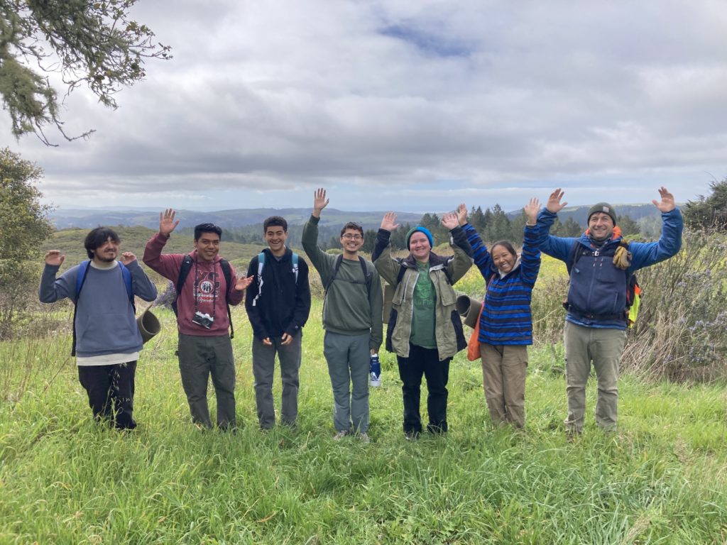 Seven people stand outside on a wide green spread of grass. There are clouds in the sky above them and they are all smiling and raising their hands.