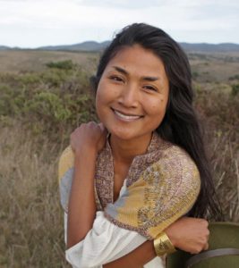 A person with long dark brown hair, wearing a golden scarf and gold bracelet, smiles at the camera. They are in front of a field of brown and green grasses with shadowed hills in the background. 
