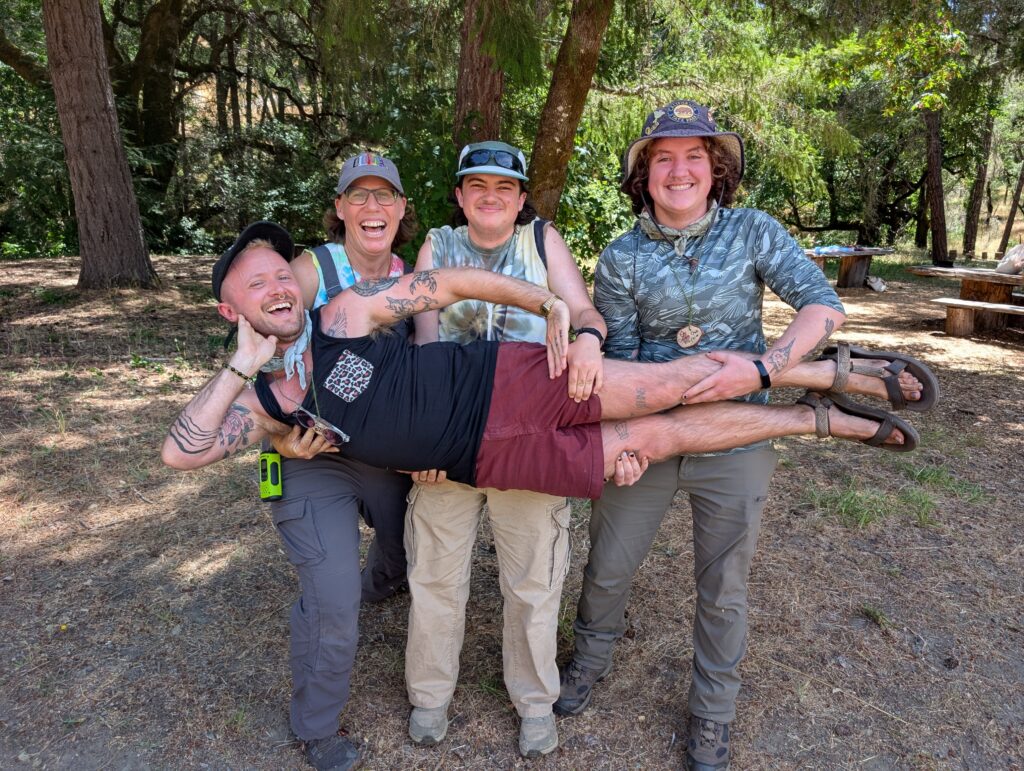 Three people are smiling as they hold up a forth person in front of a forest.
