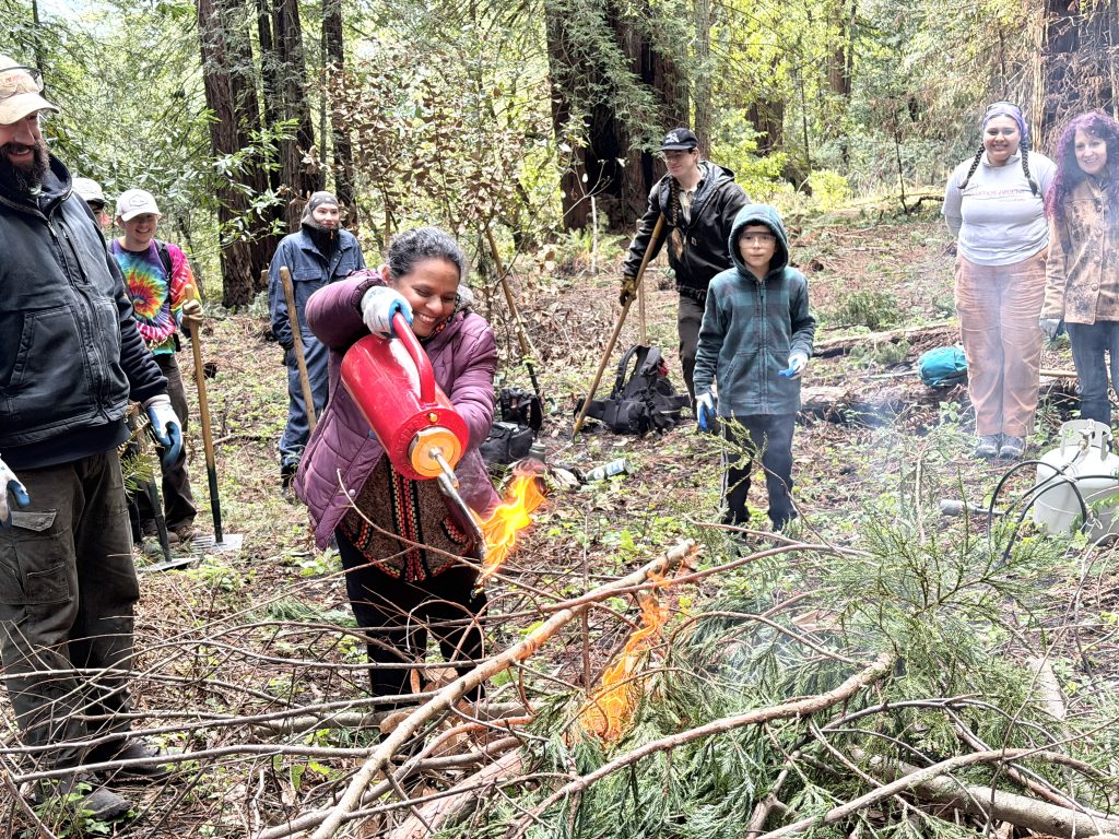 a volunteer holds a red canister and begins to start a burn pile, supporte by a landpaths staff member.
