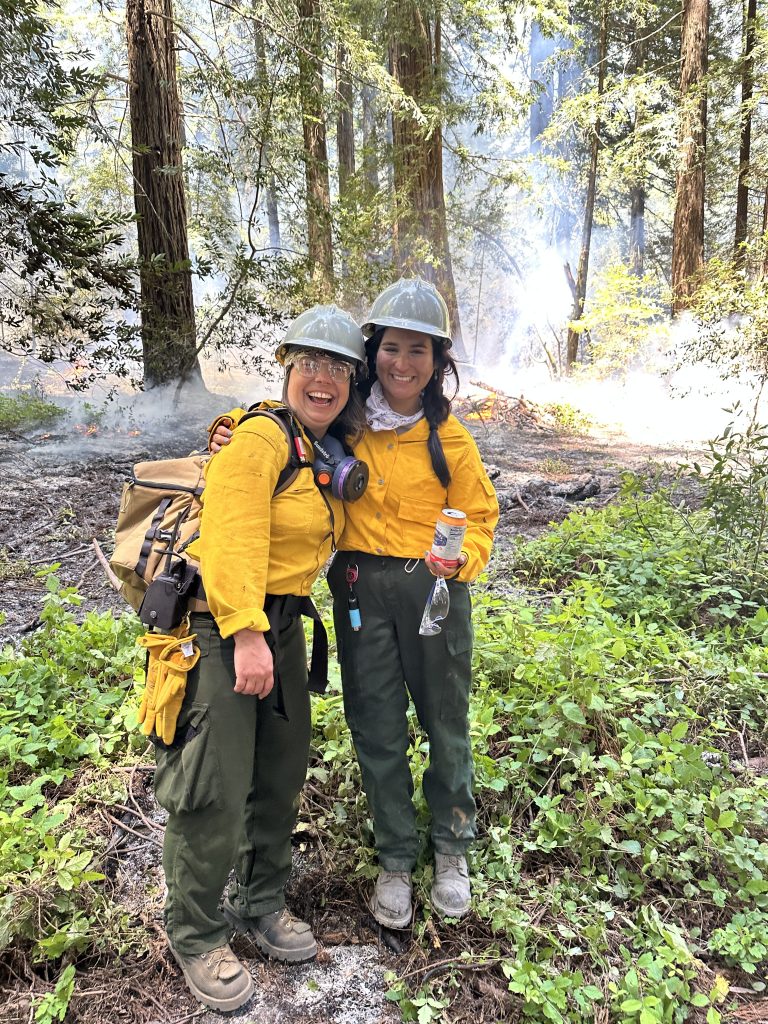Two people stand in front of a redwood grove. They are smiling and wear all of the appropriate personal protective equipment for a prescribed burn.